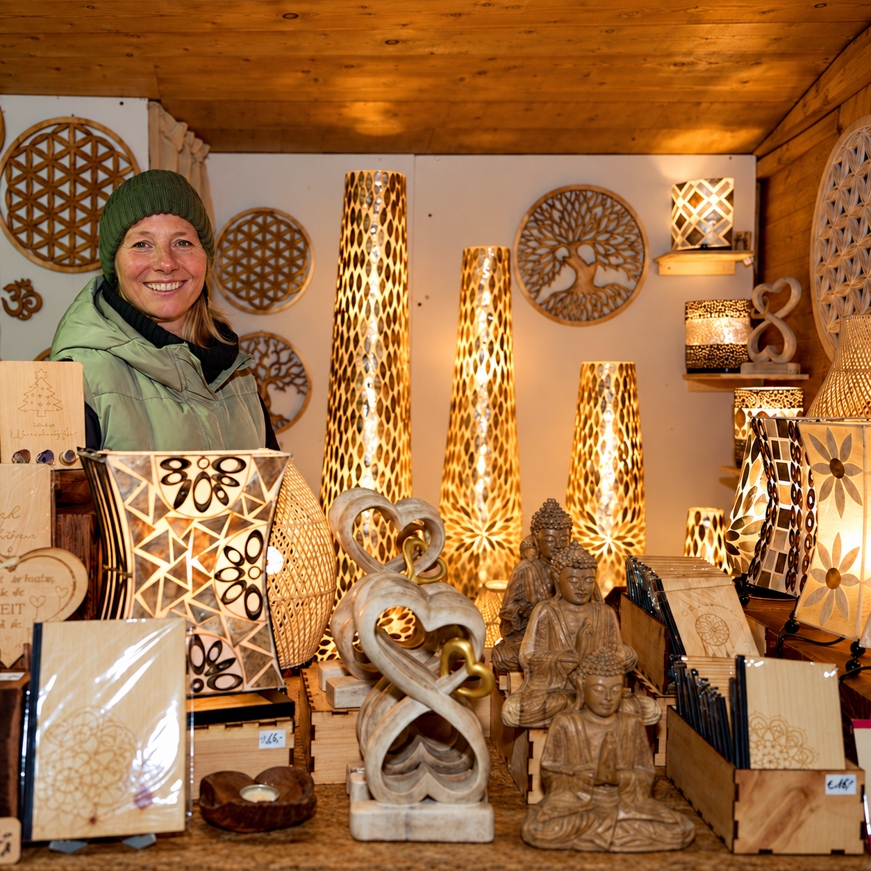 Frau am Verkaufsstand mit warm leuchtenden Holzlampen, Holzskulpturen und Karten.